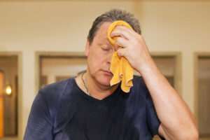 A man in a dark blue shirt wipes sweat from his forehead with an orange towel. He appears to be in a warm indoor setting.