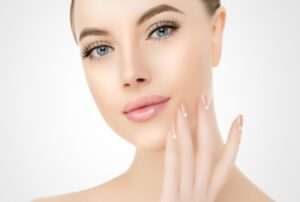 A woman with light skin, blue eyes, and glossy pink lips is touching her face with her right hand. Her nails are manicured with a nude polish and white heart designs. The background is a smooth, light gradient.