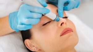 A person with closed eyes is receiving a facial treatment. Gloved hands hold a skincare device applied to their nose. The person is lying down on a white surface, appearing relaxed and calm.